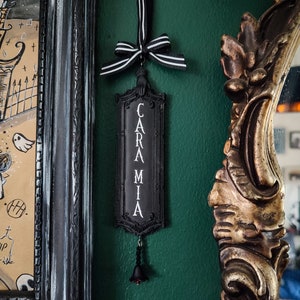 May include: Black and white sign with the word "CARAMIA" written vertically in white letters. The sign is hanging from a black and white ribbon with a black bow. A small black bell hangs from the bottom of the sign.