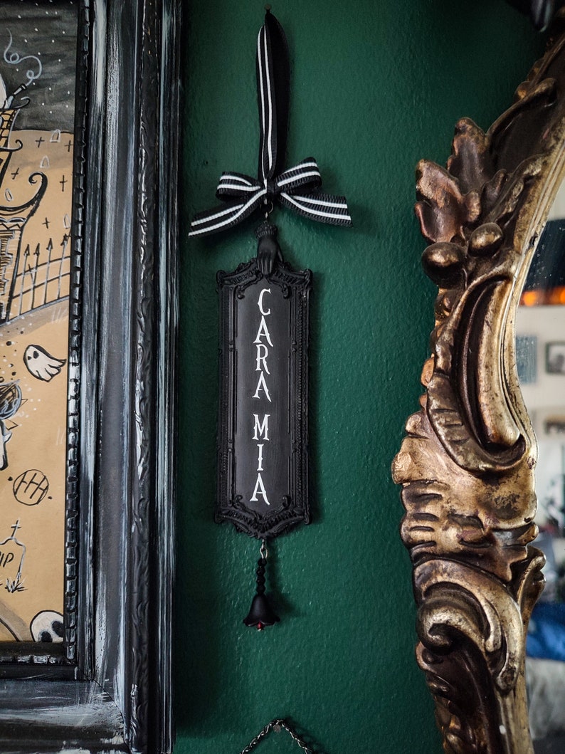May include: Black and white sign with the word "CARAMIA" written vertically in white letters. The sign is hanging from a black and white ribbon with a black bow. A small black bell hangs from the bottom of the sign.