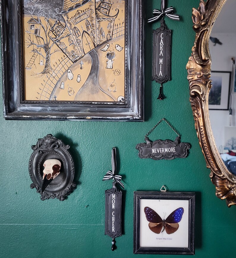 May include: A green wall with various black framed artwork and decorative items. The artwork includes a black and white drawing of a spooky house, a framed butterfly specimen, and a framed skull. The decorative items include black and white signs with the words "Nevermore", "Caramia", and "Mon Cheer".