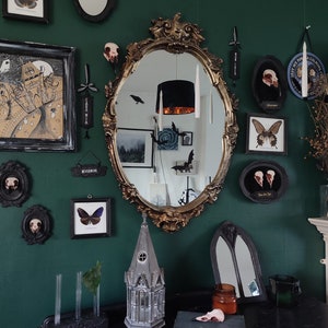 May include: A dark green room decorated with a gothic theme. The room features a large ornate gold mirror, black framed pictures, and a small model of a church. The walls are decorated with various black and white framed pictures, including a butterfly, a skull, and a bird. The room also features a small table with a book and a glass jar.