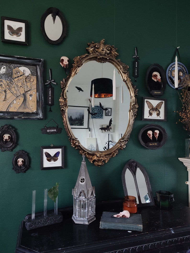 May include: A dark green room decorated with a gothic theme. The room features a large ornate gold mirror, black framed pictures, and a small model of a church. The walls are decorated with various black and white framed pictures, including a butterfly, a skull, and a bird. The room also features a small table with a book and a glass jar.