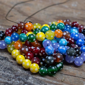May include: A selection of multi-coloured glass beads, each with white polka dots. The beads are in shades of red, yellow, blue, green, and orange. They are strung on a thin, dark cord.