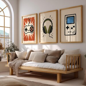 May include: Three framed art prints with gaming-themed illustrations hang above a wooden sofa. The prints feature a controller, headphones, and a handheld game console, all in a minimalist style with neutral colors and wooden frames.