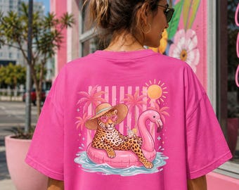 Puede incluir: Una camiseta de manga corta de color rosa brillante con un gráfico en la espalda de un leopardo con sombrero de paja y gafas de sol en forma de corazón, descansando en un flotador de flamenco rosa con palmeras, un sol amarillo y una bebida.