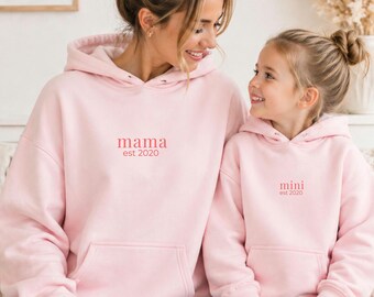 May include: Two matching light pink hoodies, one for "mama" and one for "mini", both with "est 2020" in a soft pink font. The hoodies have a front pocket and a hood. The image shows a mother and child wearing the hoodies.