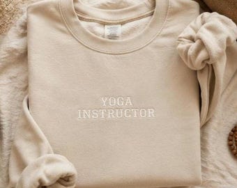 Puede incluir: Una sudadera beige de cuello redondo con las palabras YOGA INSTRUCTOR bordadas en un hilo de color claro a juego en el centro del pecho. La prenda tiene mangas largas con puños acanalados y un cuello redondo, confeccionada en tela suave.