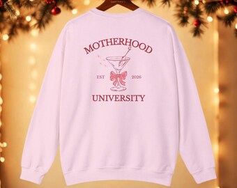 May include: A light pink sweatshirt with a graphic on the back that reads "Motherhood University" in a serif font. The graphic includes a martini glass with a bow and the year "2026". The sweatshirt is hanging on a wooden hanger.