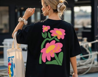 Puede incluir: Una camiseta negra con un gráfico grande en la espalda que muestra tres flores rosas estilizadas con centros amarillos y tallos verdes. El texto "egalité" está escrito en cursiva amarilla sobre los pétalos. Una bolsa de tela beige con salpicaduras de pintura.
