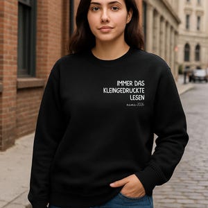 Op de afbeelding: Zwart sweatshirt met ronde hals en witte tekst die luidt: "IMMER DAS KLEINGEDRUCKTE LESEN mama 2026". Het sweatshirt wordt gedragen met een blauwe spijkerbroek. De achtergrond is een bakstenen gebouw en een geplaveide straat.