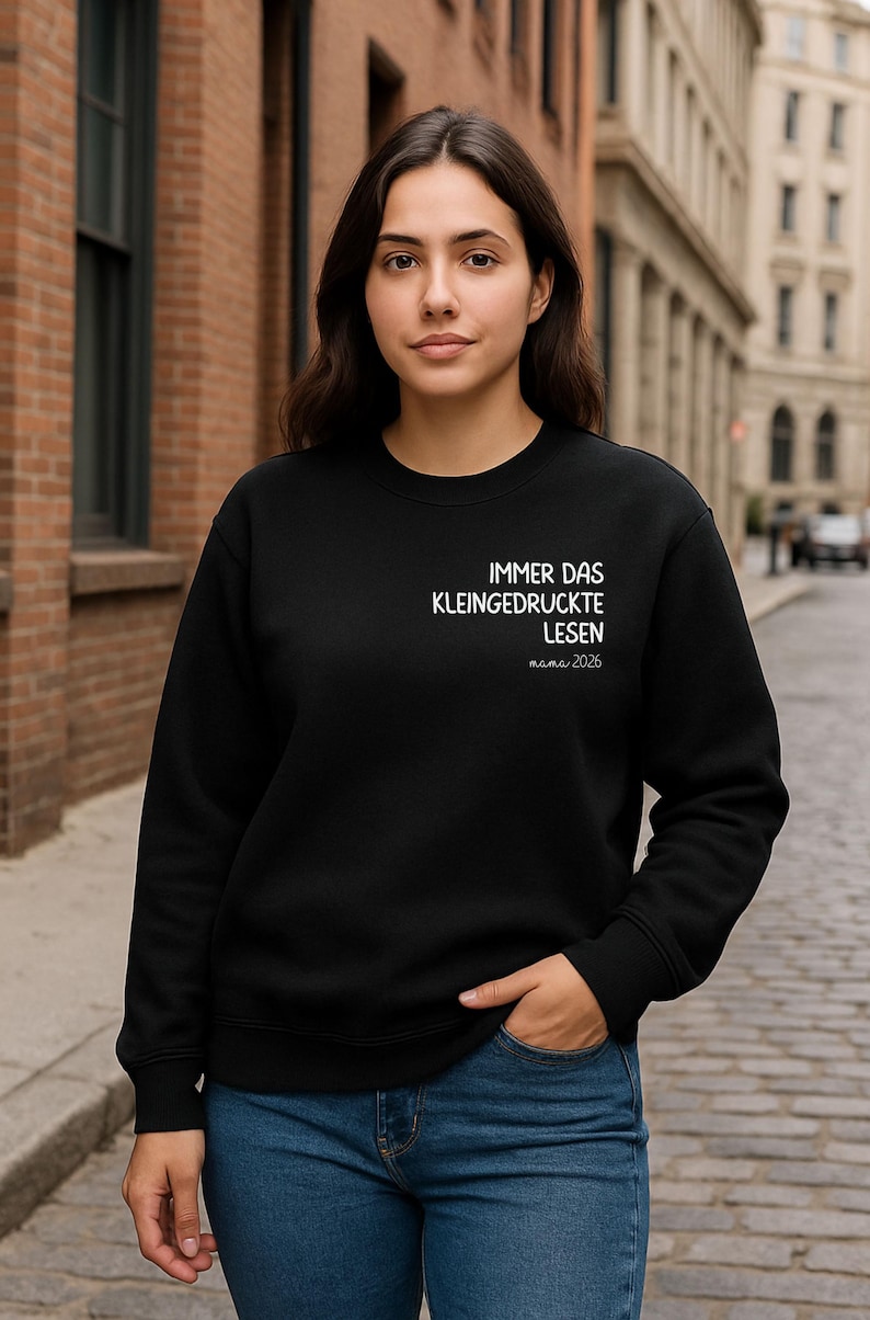 Op de afbeelding: Zwart sweatshirt met ronde hals en witte tekst die luidt: "IMMER DAS KLEINGEDRUCKTE LESEN mama 2026". Het sweatshirt wordt gedragen met een blauwe spijkerbroek. De achtergrond is een bakstenen gebouw en een geplaveide straat.