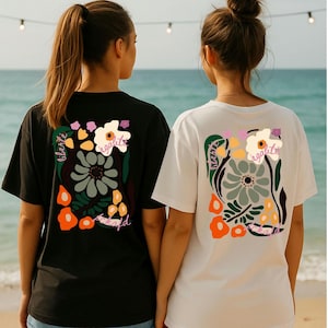 Könnte beinhalten: Zwei T-Shirts, eines schwarz und eines weiß, mit einem Blumendesign auf dem Rücken. Das Design umfasst Blumen in Orange, Grün und Weiß, mit den Wörtern "liberte" und "egalite" im Design. Die Shirts sind kurzärmelig.