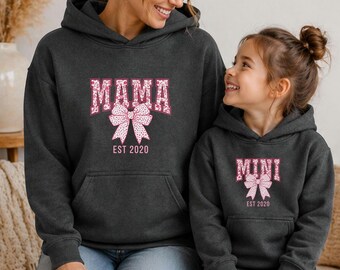 May include: Two matching charcoal gray hoodies, one adult and one child-sized, featuring the words "MAMA" and "MINI" in a floral font with a bow graphic. The hoodies also have "EST 2020" printed below the bow.