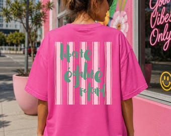 Puede incluir: Una camiseta de manga corta de color rosa brillante con un gráfico en la espalda de rayas verticales de color rosa claro y el texto verde "liberté égalité fraternité" en una fuente redondeada. La prenda tiene cuello redondo y corte holgado.