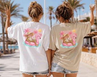 Puede incluir: Una camiseta blanca y una camiseta beige, cada una con un gráfico en la espalda de un leopardo con sombrero de paja y gafas de sol, descansando sobre un flotador de flamenco rosa. El diseño incluye palmeras rosas, un sol amarillo y olas.