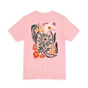Puede incluir: Una camiseta rosa claro con un dise&ntilde;o floral. El dise&ntilde;o incluye las palabras "liberte" y "egalite" en un estilo manuscrito, junto con flores en verde, naranja y blanco.