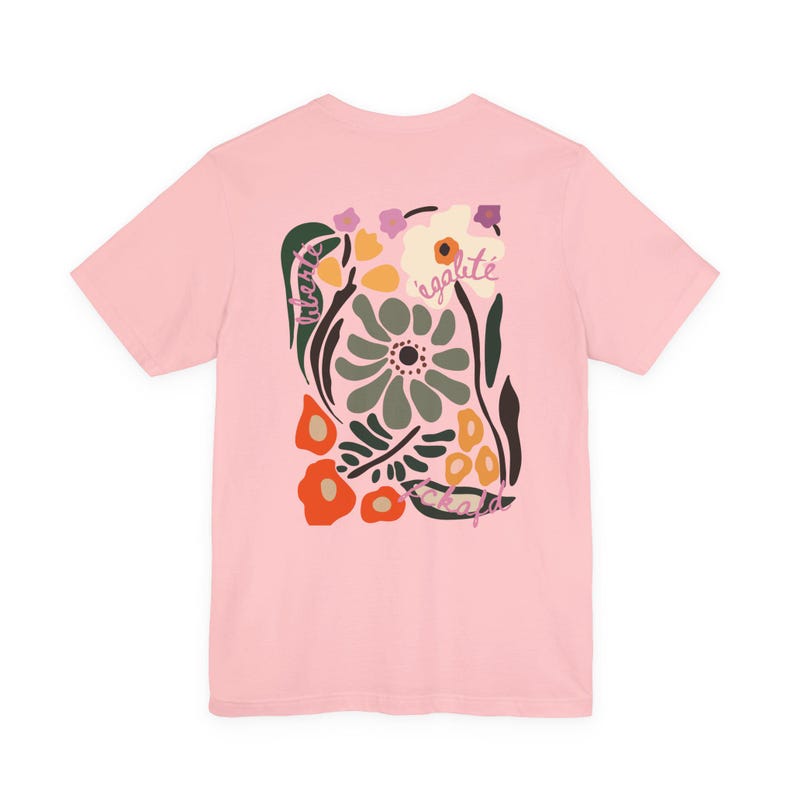 Puede incluir: Una camiseta rosa claro con un dise&ntilde;o floral. El dise&ntilde;o incluye las palabras "liberte" y "egalite" en un estilo manuscrito, junto con flores en verde, naranja y blanco.