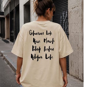 Könnte beinhalten: Ein beiges Kurzarm-T-Shirt mit schwarzem Text auf der Rückseite. Der Text ist in zwei Spalten angeordnet und lautet: Geburtsort Erde, Rasse Mensch, Politik Freiheit, Religion Liebe. Das T-Shirt hat einen Rundhalsausschnitt und eine lockere Passform.