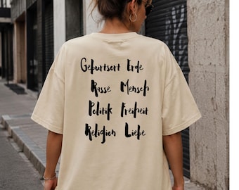 Puede incluir: Una camiseta beige de manga corta con texto negro impreso en la parte posterior. El texto está dispuesto en dos columnas y dice: Geburtsort Erde, Rasse Mensch, Politik Freiheit, Religion Liebe. La camiseta tiene cuello redondo y corte holgado.