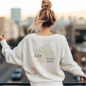 May include: A cream-colored sweatshirt with the words "GO Sage YOUR SELF" printed on the back in a mix of fonts and colors. The design includes a sage-green illustration. The person is standing outside with a city view in the background.