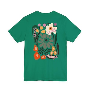 Puede incluir: Una camiseta verde con un dise&ntilde;o floral. El dise&ntilde;o incluye las palabras "libert&eacute;, &eacute;galit&eacute;, fraternit&eacute;" en rosa y blanco, y flores naranjas, rosas y blancas, y una gran flor marr&oacute;n.