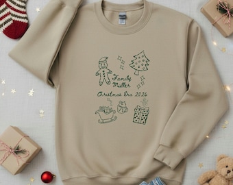 May include: A beige crewneck sweatshirt featuring dark green line art illustrations of a gingerbread man, a Christmas tree, a sleigh, a mug, and a gift box, with the text "Family Müller Christmas Era 2026" printed in the center.