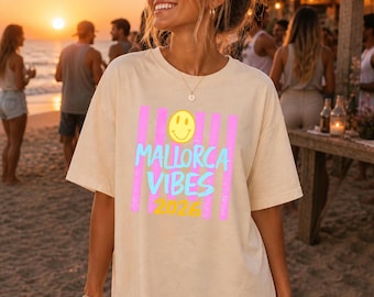 May include: Beige t-shirt with the text "MALLORCA VIBES 2026" in pink, blue, and yellow. The shirt features a smiley face graphic and vertical stripes. The person is holding a drink with a straw.
