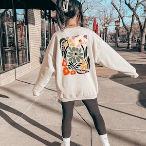 May include: Cream-colored sweatshirt with a floral graphic on the back, featuring orange, green, and white flowers, and the word "&eacute;galit&eacute;". The sweatshirt is long-sleeved and worn with black leggings and white sneakers.