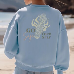 May include: Light blue sweatshirt with the text "Go Sage Yourself" and a sage bundle graphic on the back. The design features a sage bundle with flames and text in a mix of script and block letters. The sweatshirt is a relaxed fit.