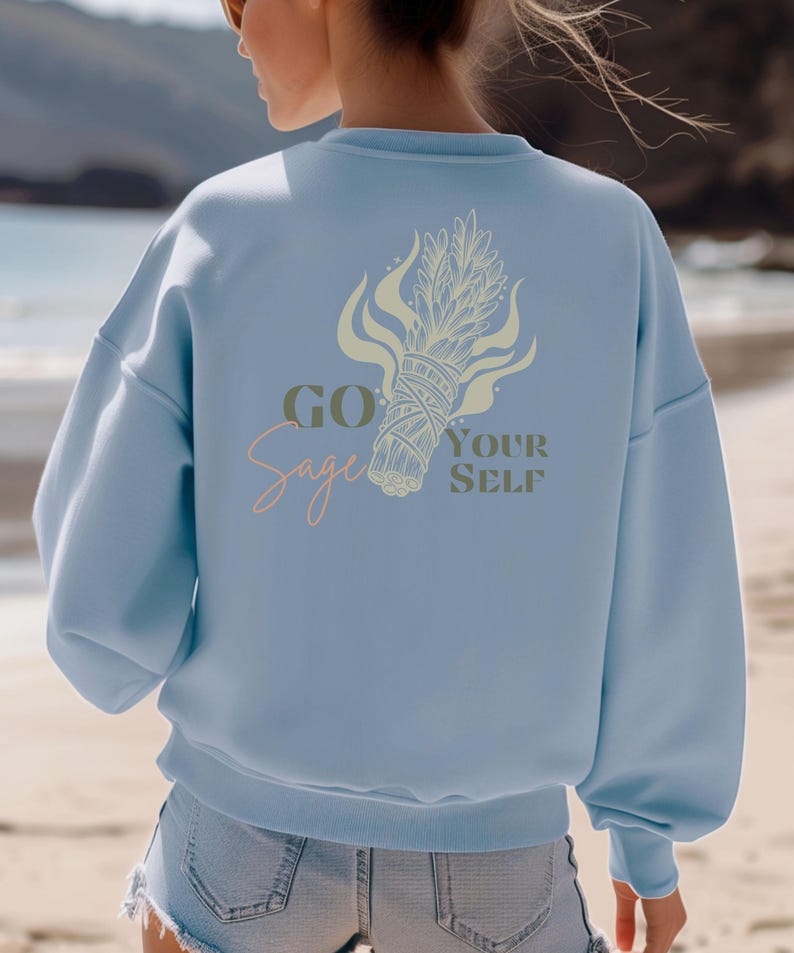 May include: Light blue sweatshirt with the text "Go Sage Yourself" and a sage bundle graphic on the back. The design features a sage bundle with flames and text in a mix of script and block letters. The sweatshirt is a relaxed fit.