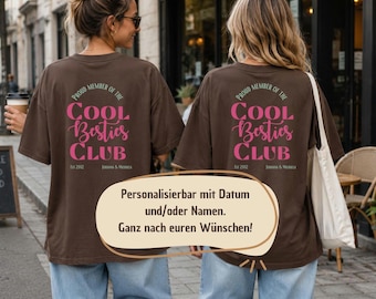 May include: A brown short-sleeved t-shirt featuring the text "PROUD MEMBER OF THE COOL BESTIES CLUB" in pink and green, with "EST 2012" and "JOHANNA & MICHAELA" printed below. The design is centered on the back of the shirt.
