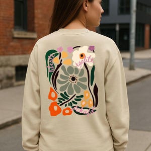 May include: Beige sweatshirt with a colorful floral design on the back. The design includes flowers in shades of green, orange, and purple, with the word "egalite" in cursive.