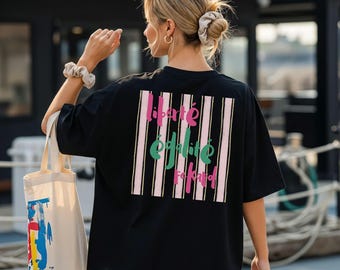 Puede incluir: Una camiseta negra de manga corta con un gráfico rectangular en la espalda con rayas verticales rosas y blancas. El texto "liberté", "égalité" y "fraternité" está impreso en letra cursiva rosa y verde. Se lleva una bolsa de tela de color beige.