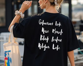 Puede incluir: Una camiseta negra de manga corta con texto blanco escrito a mano en la espalda que dice Geburtsort Erde, Rasse Mensch, Politik Freiheit, Religion Liebe. La camiseta se combina con una bolsa de tela beige con un estampado abstracto colorido.