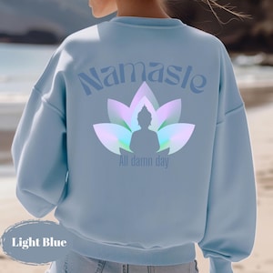 May include: Light blue sweatshirt with the word "Namaste" above a lotus flower and Buddha silhouette. The text "All damn day" is below the Buddha. The sweatshirt is light blue.