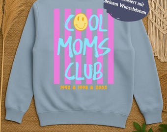 Puede incluir: Sudadera azul claro con las palabras "COOL MOMS CLUB" en turquesa, una carita sonriente amarilla y rayas verticales rosas. La sudadera también tiene los años 1992, 1998 y 2003.