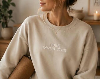 Puede incluir: Una sudadera beige de cuello redondo con el texto YOGA INSTRUCTOR bordado en hilo blanco en el pecho. La prenda tiene mangas largas, un cuello acanalado y un corte holgado, ofreciendo un tono neutro y un diseño sencillo.