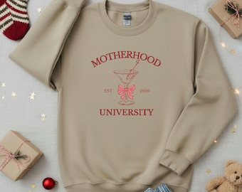May include: A beige sweatshirt with the words "MOTHERHOOD UNIVERSITY" in red script. A martini glass graphic with a pink bow is centered below the word "MOTHERHOOD". The year "2026" is printed on the right side of the graphic.