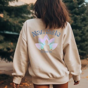 May include: Beige sweatshirt with the word "Namaste" in light blue script above a lotus flower design with a silhouette of a person in the center. The text "All damn day" is below the lotus flower.