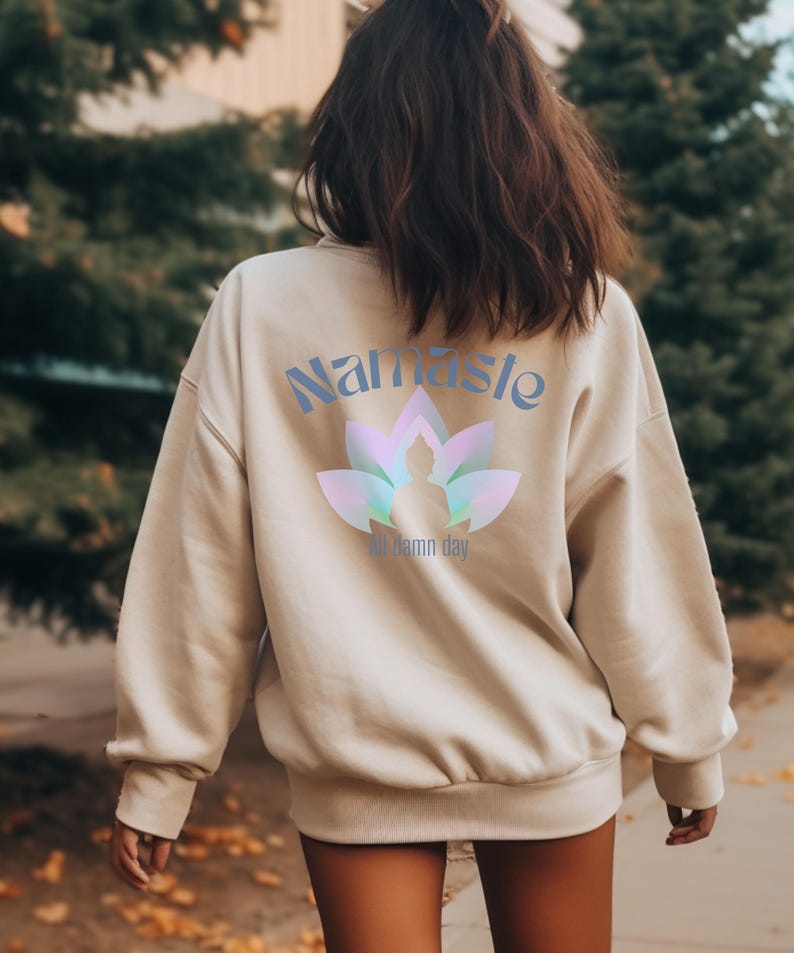 May include: Beige sweatshirt with the word "Namaste" in light blue script above a lotus flower design with a silhouette of a person in the center. The text "All damn day" is below the lotus flower.