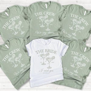 Könnte beinhalten: Eine Auswahl an grünen und weißen T-Shirts. Die grünen Shirts zeigen "THE CREW" mit einem Cocktail-Design und "We say cheers". Das weiße Shirt trägt die Aufschrift "THE BRIDE" mit einer Cocktail-Grafik und "I said yes".