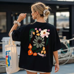 Könnte beinhalten: Schwarzes T-Shirt mit floralem Design auf dem Rücken, mit den Worten "liberte, egalite, fraternite". Das Shirt wird mit einer cremefarbenen Tragetasche mit farbenfrohem, abstraktem Design kombiniert.