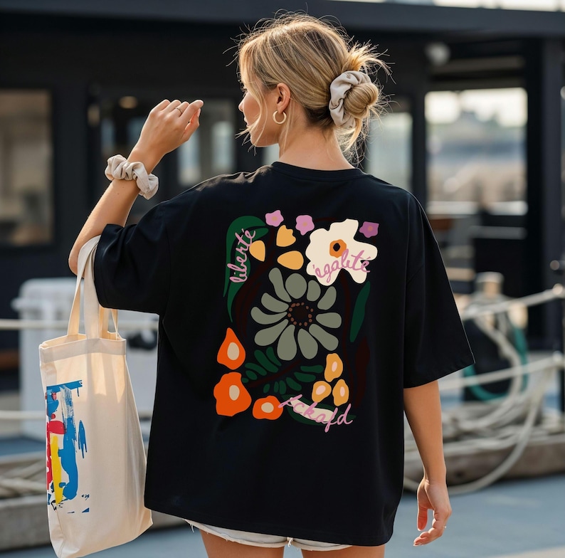 Puede incluir: Camiseta negra con un dise&ntilde;o floral en la espalda, con las palabras "liberte, egalite, fraternite". La camiseta se combina con una bolsa de mano color crema con un dise&ntilde;o abstracto colorido.