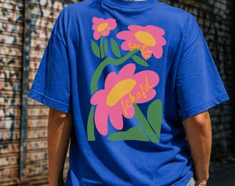 Puede incluir: Una camiseta de manga corta color azul real con un gráfico grande en la espalda. El diseño muestra tres flores rosas con centros amarillos y tallos verdes. Las palabras "liberté", "égalité" y "fckafd" están escritas en cursiva amarilla.