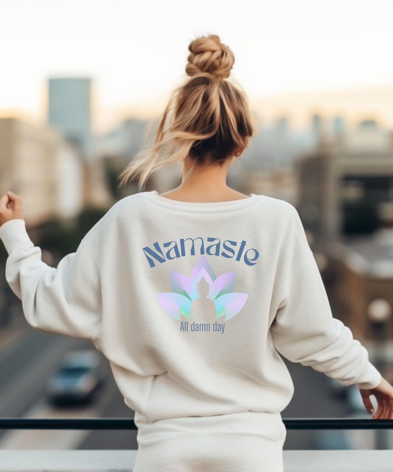May include: Cream-colored sweatshirt with the word "Namaste" in blue script above a lotus flower design with a person in the center. Below the lotus flower is the text "All damn day". The sweatshirt is a casual style.
