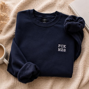 Puede incluir: Una sudadera azul marino de cuello redondo con el texto FCK NZS bordado en blanco en el pecho izquierdo. La prenda tiene mangas largas con puños acanalados y un forro interior de forro polar suave con un diseño sencillo y limpio.