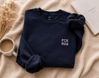 Puede incluir: Una sudadera azul marino de cuello redondo con el texto FCK NZS bordado en blanco en el pecho izquierdo. La prenda tiene mangas largas con puños acanalados y un forro interior de forro polar suave con un diseño sencillo y limpio.