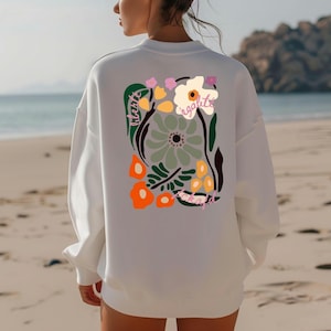 May include: White sweatshirt with a colorful floral design on the back. The design includes orange, green, pink, and white flowers with the word "egalite" in pink. The sweatshirt is a casual style, perfect for everyday wear.