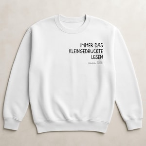 Op de afbeelding: Witte crewneck sweatshirt met zwarte tekst. De tekst luidt "IMMER DAS KLEINGEDRUCKTE LESEN mama 2026". De sweatshirt heeft lange mouwen en een ronde hals. De tekst is gecentreerd op de borst.