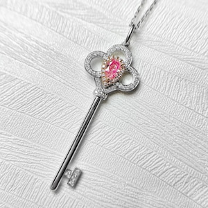 May include: A silver key pendant necklace with a pear-shaped pink gemstone at the center. The key's head is a four-leaf clover design, encrusted with small clear gemstones. The key's stem and the key's teeth are also silver.