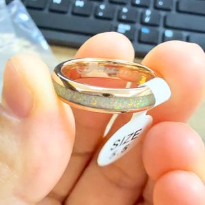 May include: A gold-colored ring with a light-colored, textured inlay. The ring is held by a person. A white tag with the word "SIZE" and the number "5.5" is attached to the ring.
